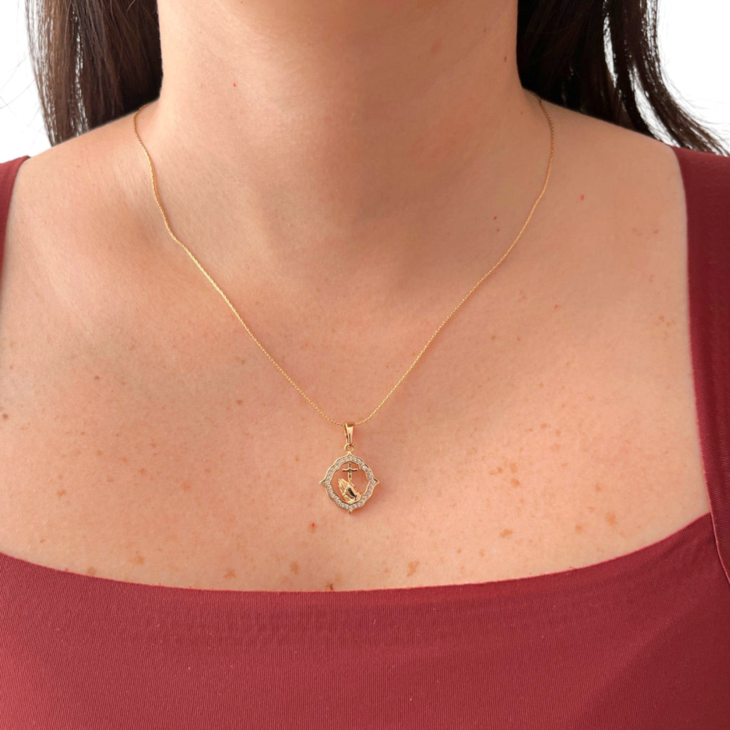 Cadena fina con tejido de estilo Anchor que lleva un dije tipo medalla de forma geométrica labrada. En el centro cuenta con el detalle de manos en posición de oración sosteniendo una cruz, rodeado por un marco engastado con circonias brillantes.