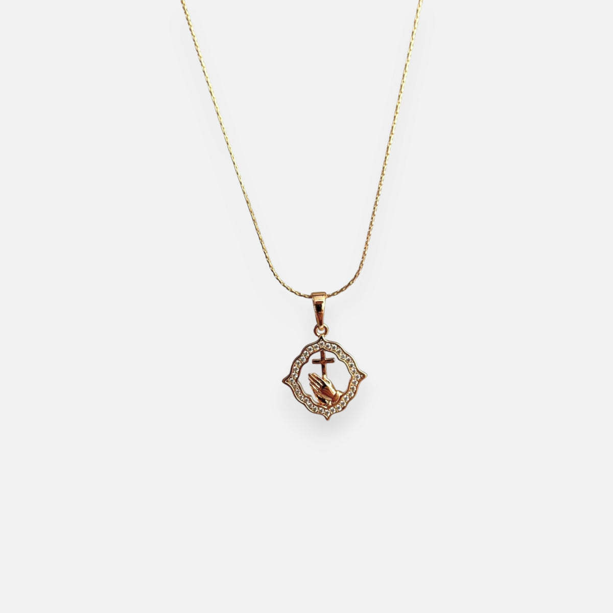 Cadena fina con tejido de estilo Anchor que lleva un dije tipo medalla de forma geométrica labrada. En el centro cuenta con el detalle de manos en posición de oración sosteniendo una cruz, rodeado por un marco engastado con circonias brillantes.