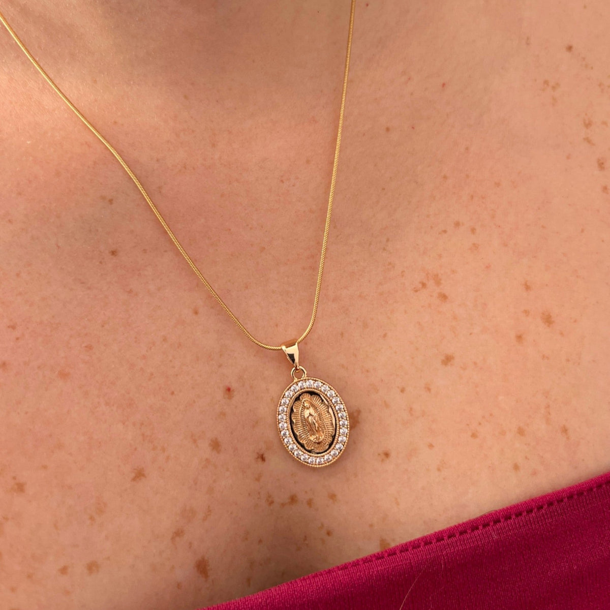 Cadena fina de tejido Cola de Ratón en acabado dorado, acompañada por un dije de medalla ovalada con la figura central de la Virgen de Guadalupe sobre relieve radiante. El contorno está delicadamente decorado con un cerco de incrustaciones de circonias brillantes.