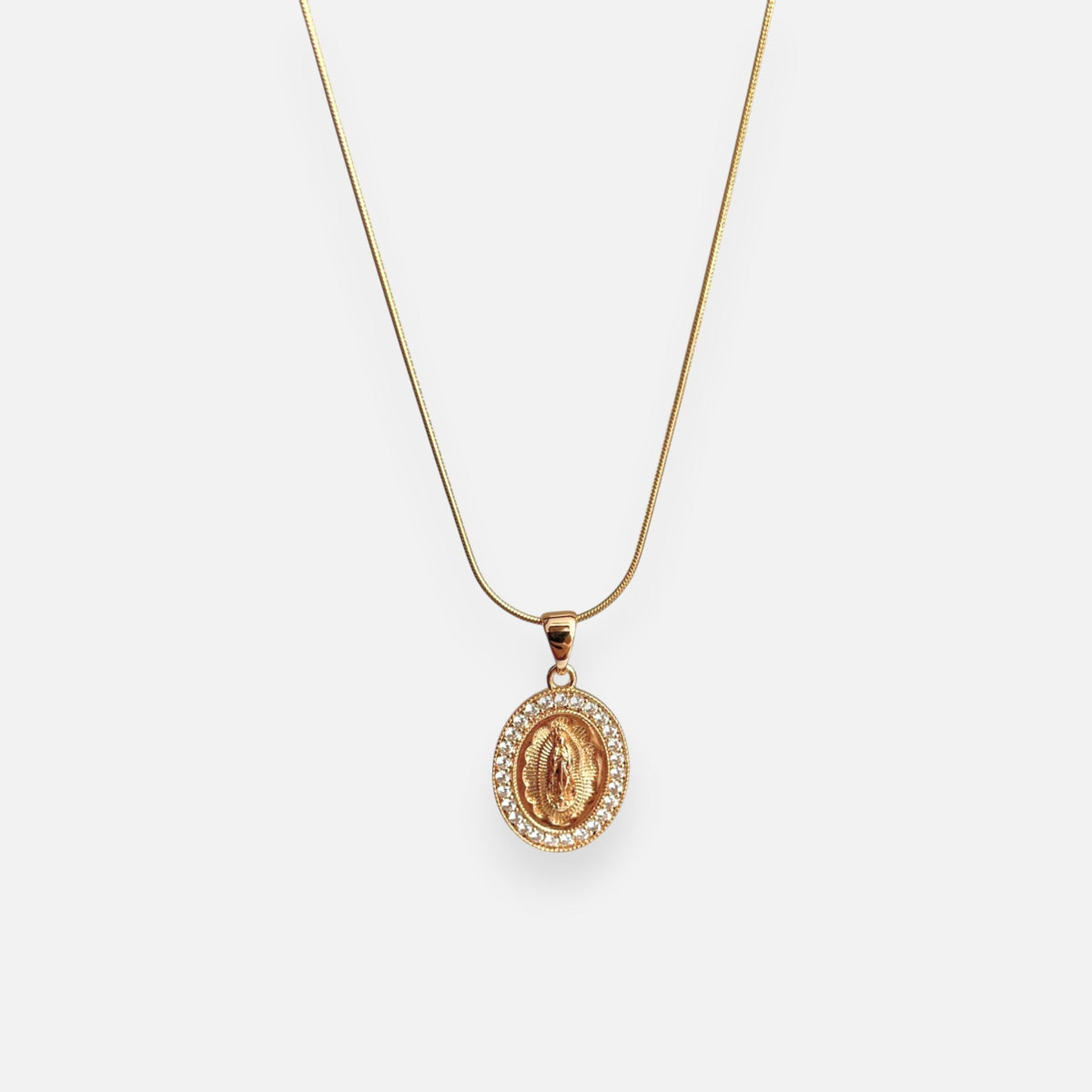 Cadena fina de tejido Cola de Ratón en acabado dorado, acompañada por un dije de medalla ovalada con la figura central de la Virgen de Guadalupe sobre relieve radiante. El contorno está delicadamente decorado con un cerco de incrustaciones de circonias brillantes.