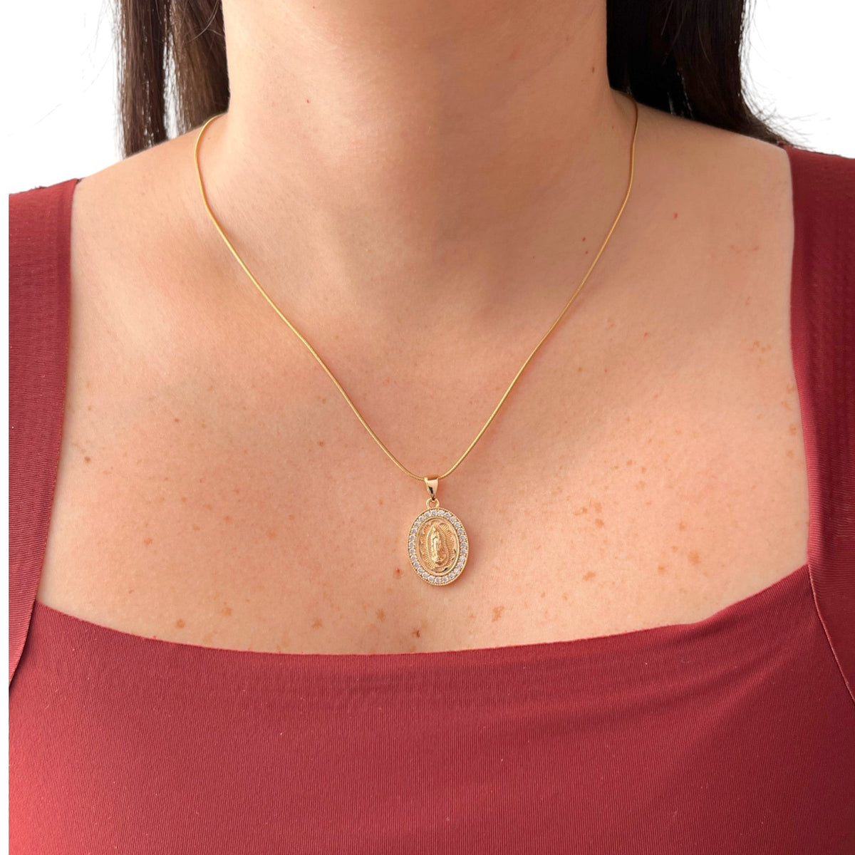 Cadena fina de tejido Cola de Ratón en acabado dorado, acompañada por un dije de medalla ovalada con la figura central de la Virgen de Guadalupe sobre relieve radiante. El contorno está delicadamente decorado con un cerco de incrustaciones de circonias brillantes.