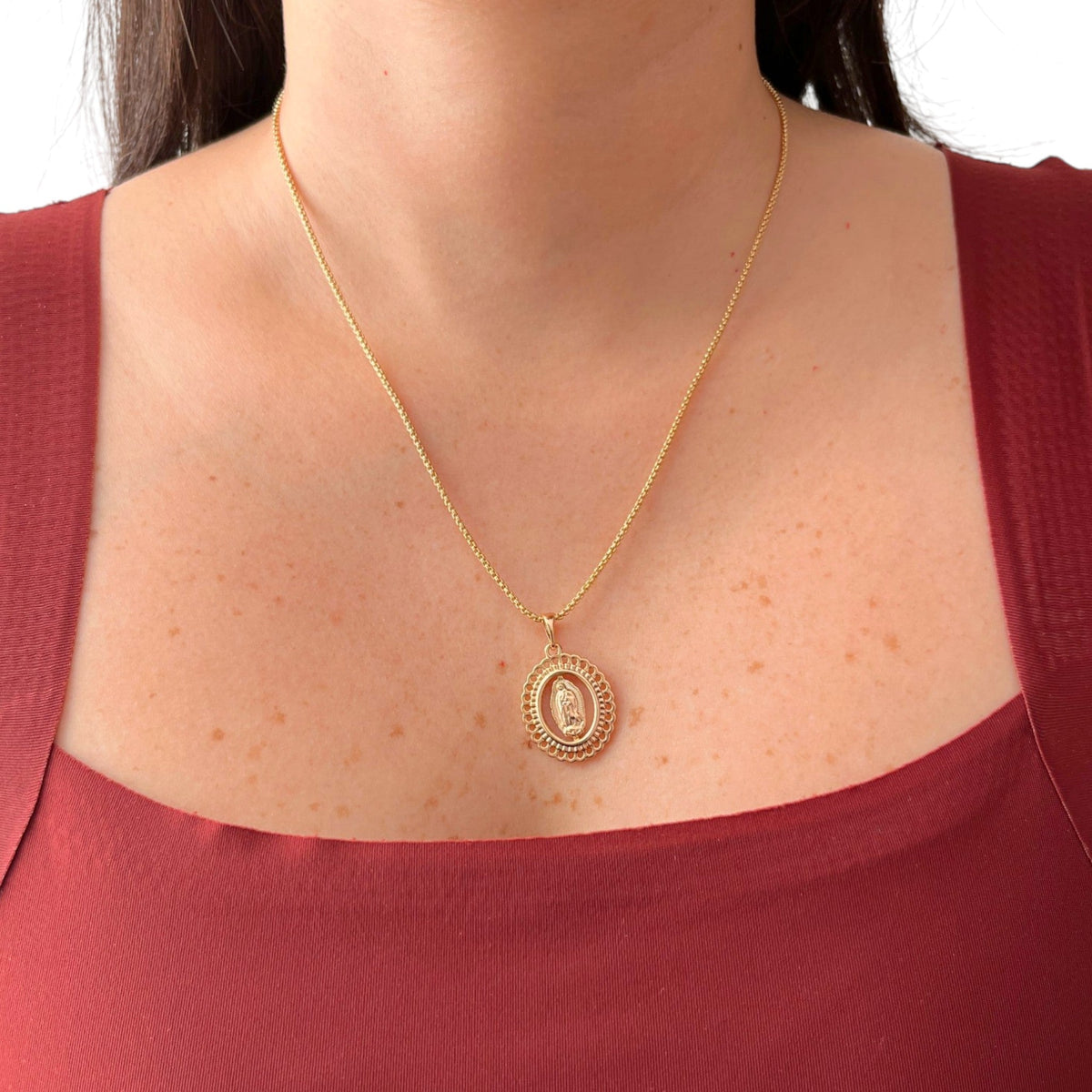 Cadena de tejido veneciana en acabado dorado, acompañada por un dije en forma de medalla ovalada con la figura central en relieve de la Virgen de Guadalupe. La pieza destaca por su elegante borde exterior calado con detalles troquelados en forma de encaje o filigrana.
