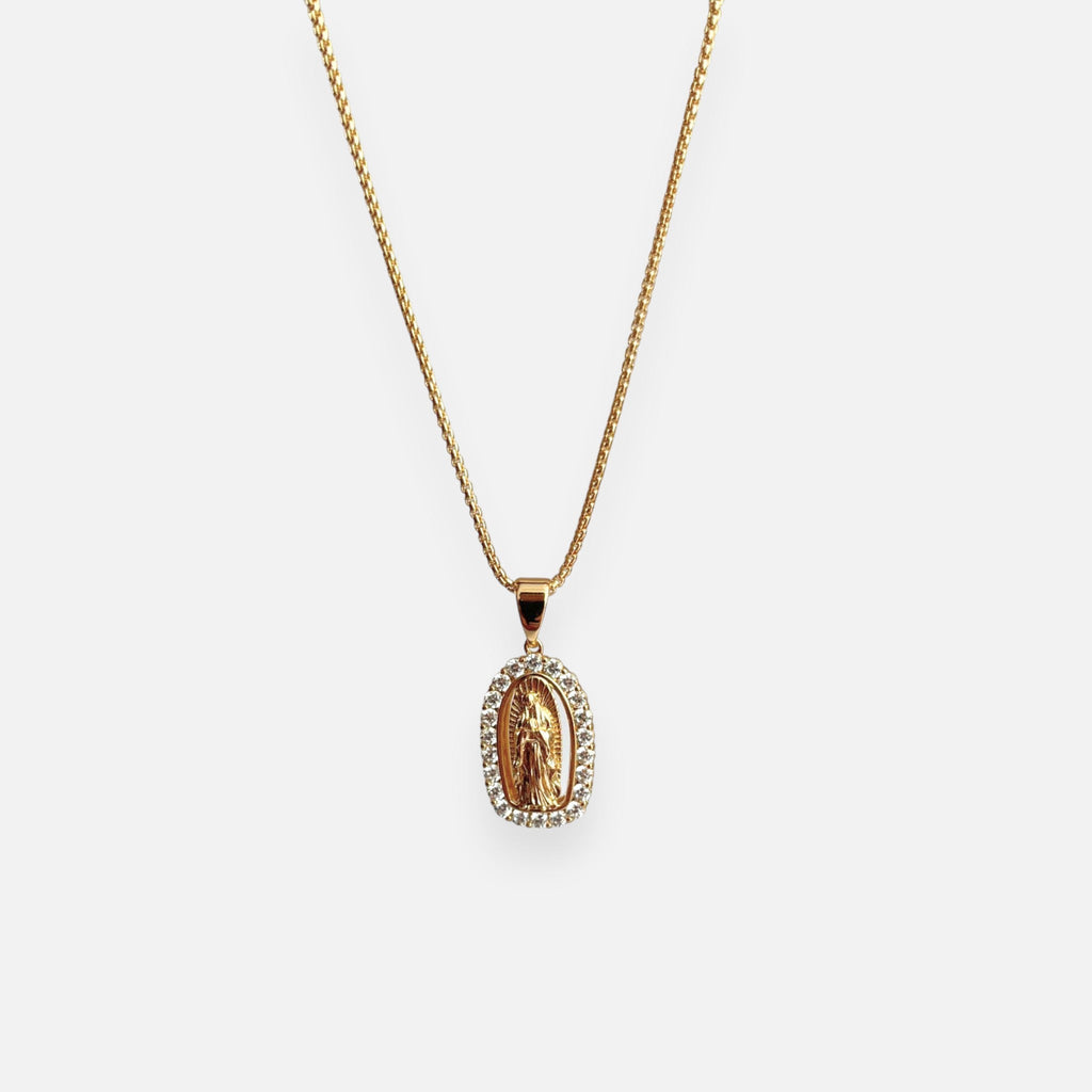 Cadena fina de tejido veneciana redondeada en acabado dorado, acompañada por un dije de medalla ovalada con la figura en relieve de la Virgen de Guadalupe. La pieza está enmarcada por un borde engastado con circonias brillantes.