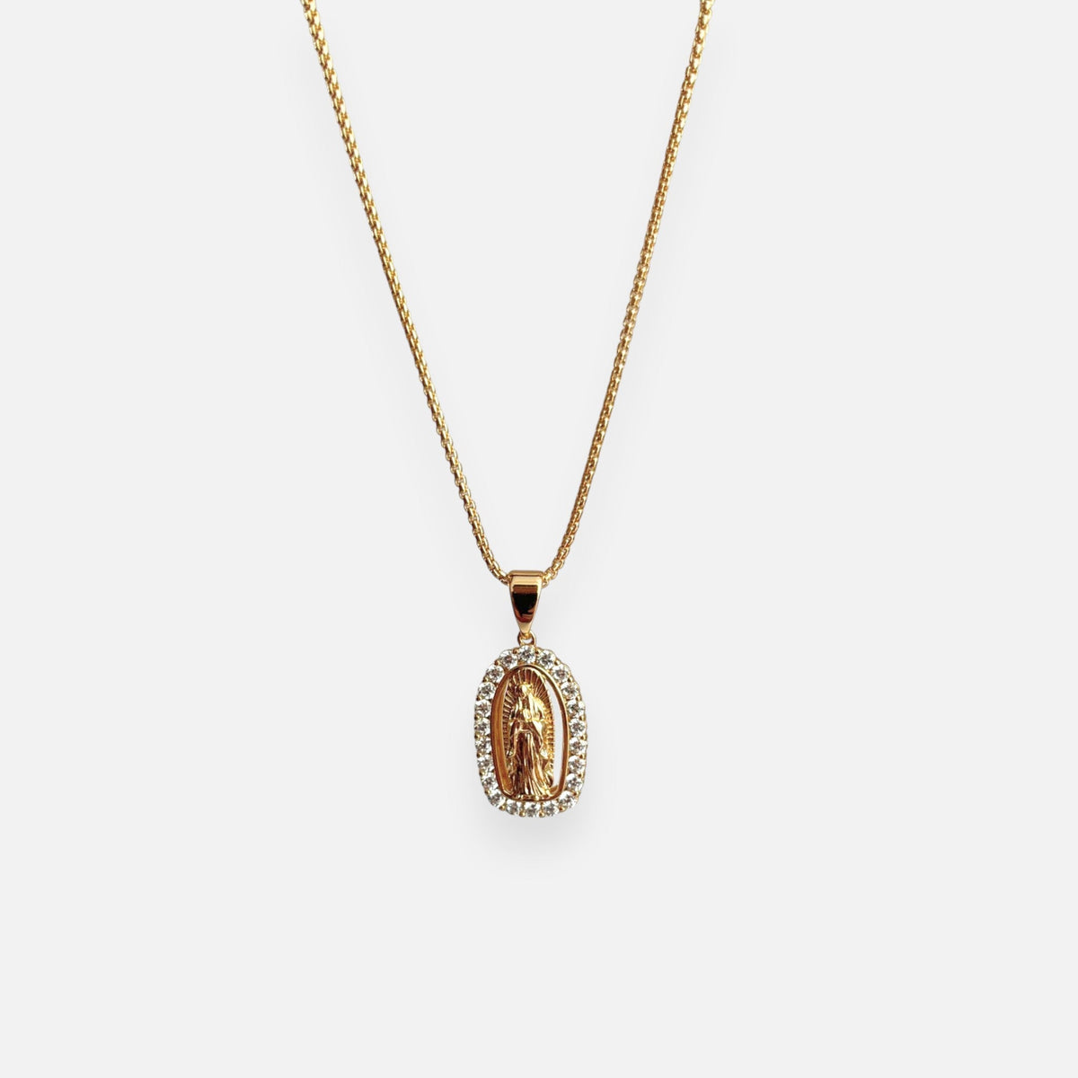 Cadena fina de tejido veneciana redondeada en acabado dorado, acompañada por un dije de medalla ovalada con la figura en relieve de la Virgen de Guadalupe. La pieza está enmarcada por un borde engastado con circonias brillantes.