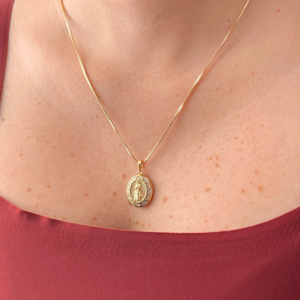 Cadena fina de tejido Veneciana en tono dorado con un dije en forma de medalla ovalada. Presenta la imagen central de la Virgen de Guadalupe sobre un fondo radiante, rodeada por un cerco engastado en incrustaciones de circonias brillantes