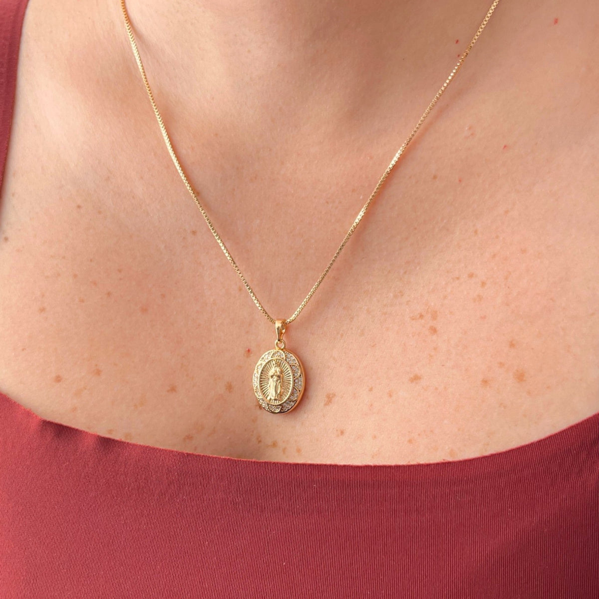 Cadena fina de tejido Veneciana en tono dorado con un dije en forma de medalla ovalada. Presenta la imagen central de la Virgen de Guadalupe sobre un fondo radiante, rodeada por un cerco engastado en incrustaciones de circonias brillantes