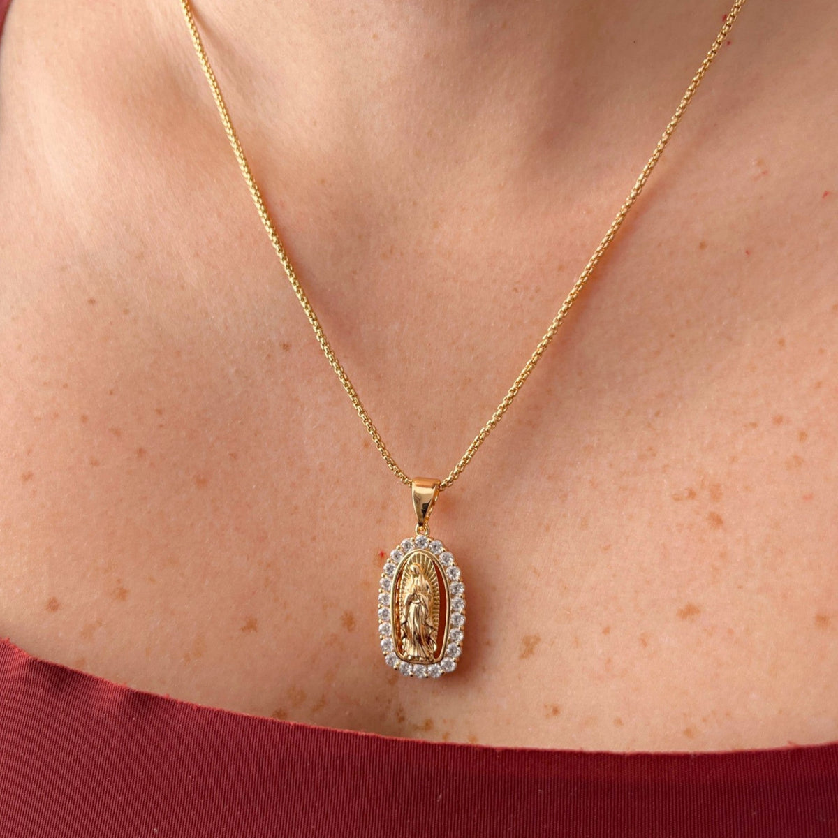 Cadena fina de tejido veneciana redondeada en acabado dorado, acompañada por un dije de medalla ovalada con la figura en relieve de la Virgen de Guadalupe. La pieza está enmarcada por un borde engastado con circonias brillantes.