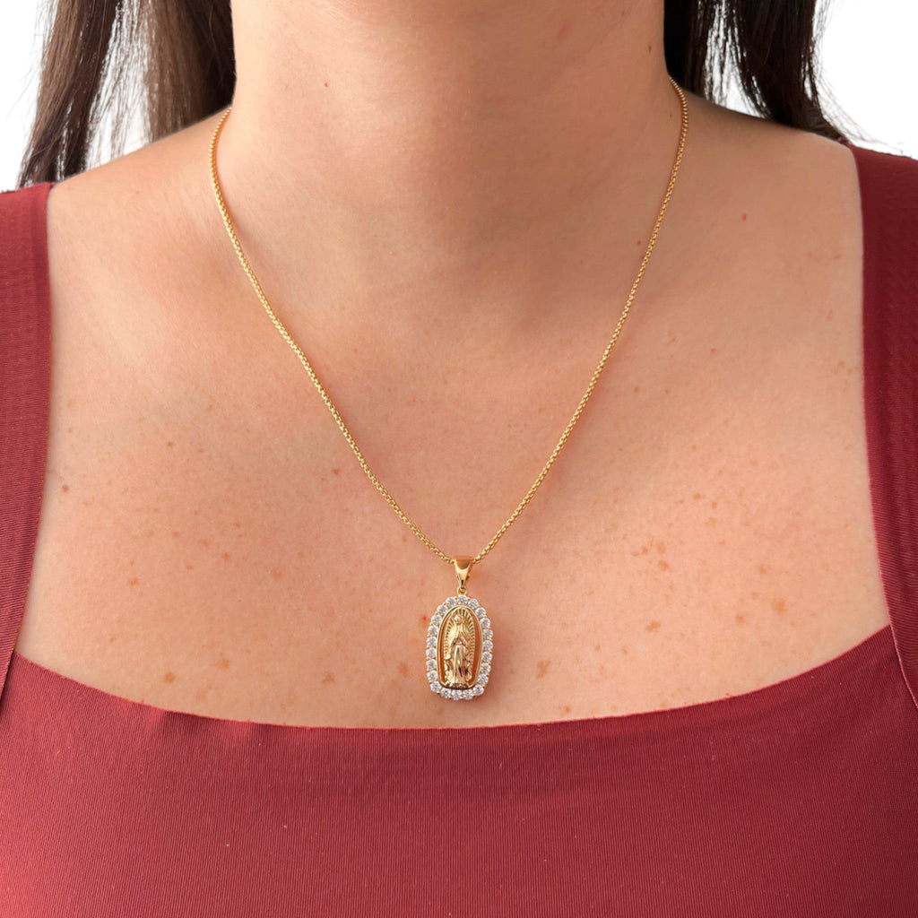 Cadena fina de tejido veneciana redondeada en acabado dorado, acompañada por un dije de medalla ovalada con la figura en relieve de la Virgen de Guadalupe. La pieza está enmarcada por un borde engastado con circonias brillantes.