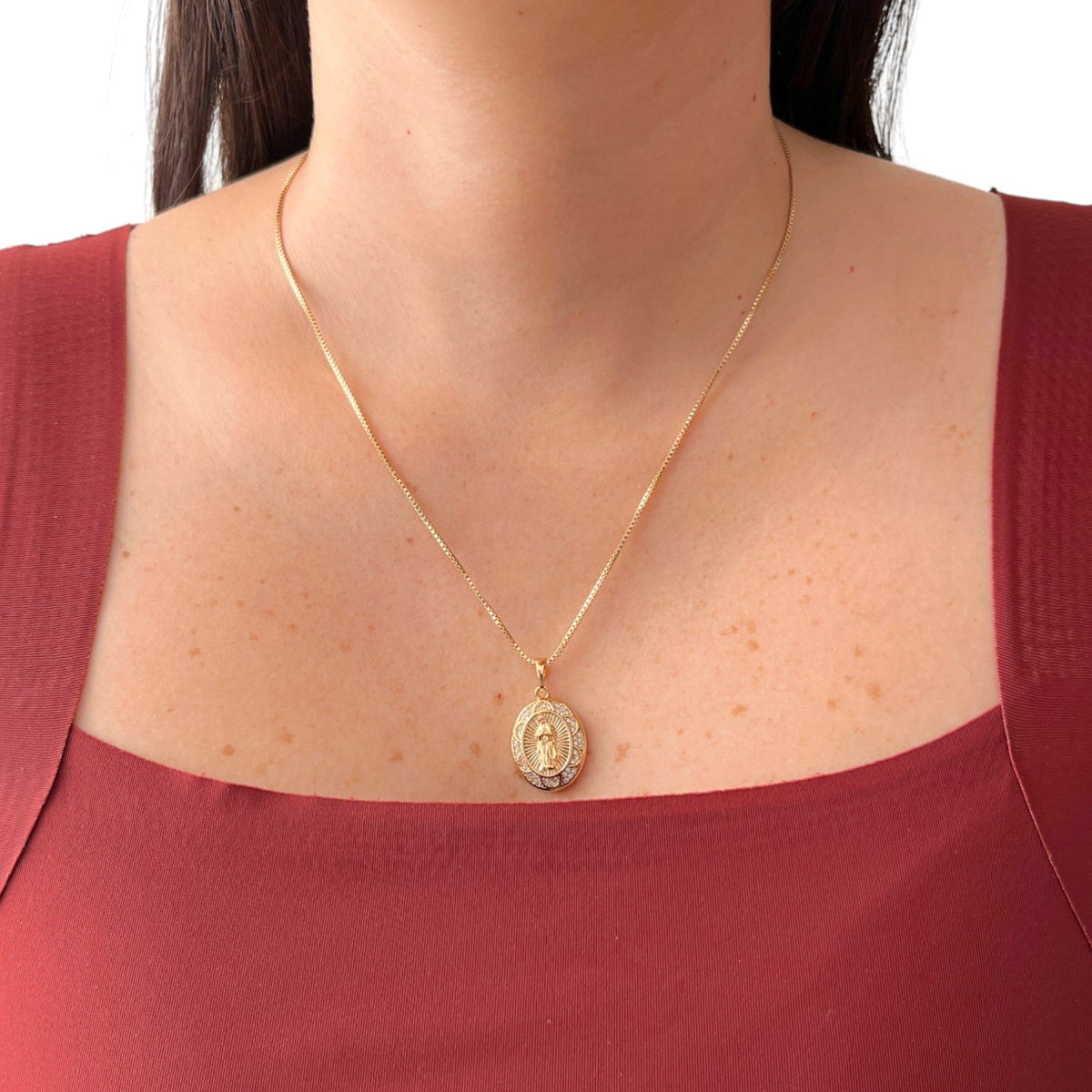 Cadena fina de tejido Veneciana en tono dorado con un dije en forma de medalla ovalada. Presenta la imagen central de la Virgen de Guadalupe sobre un fondo radiante, rodeada por un cerco engastado en incrustaciones de circonias brillantes