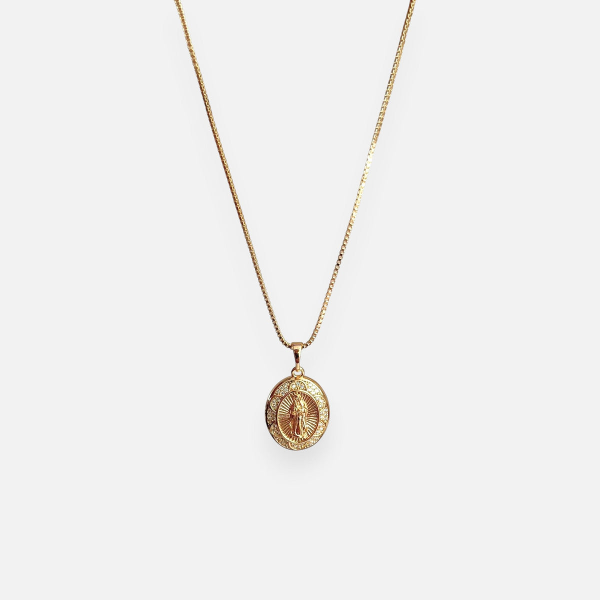 Cadena fina de tejido Veneciana en tono dorado con un dije en forma de medalla ovalada. Presenta la imagen central de la Virgen de Guadalupe sobre un fondo radiante, rodeada por un cerco engastado en incrustaciones de circonias brillantes