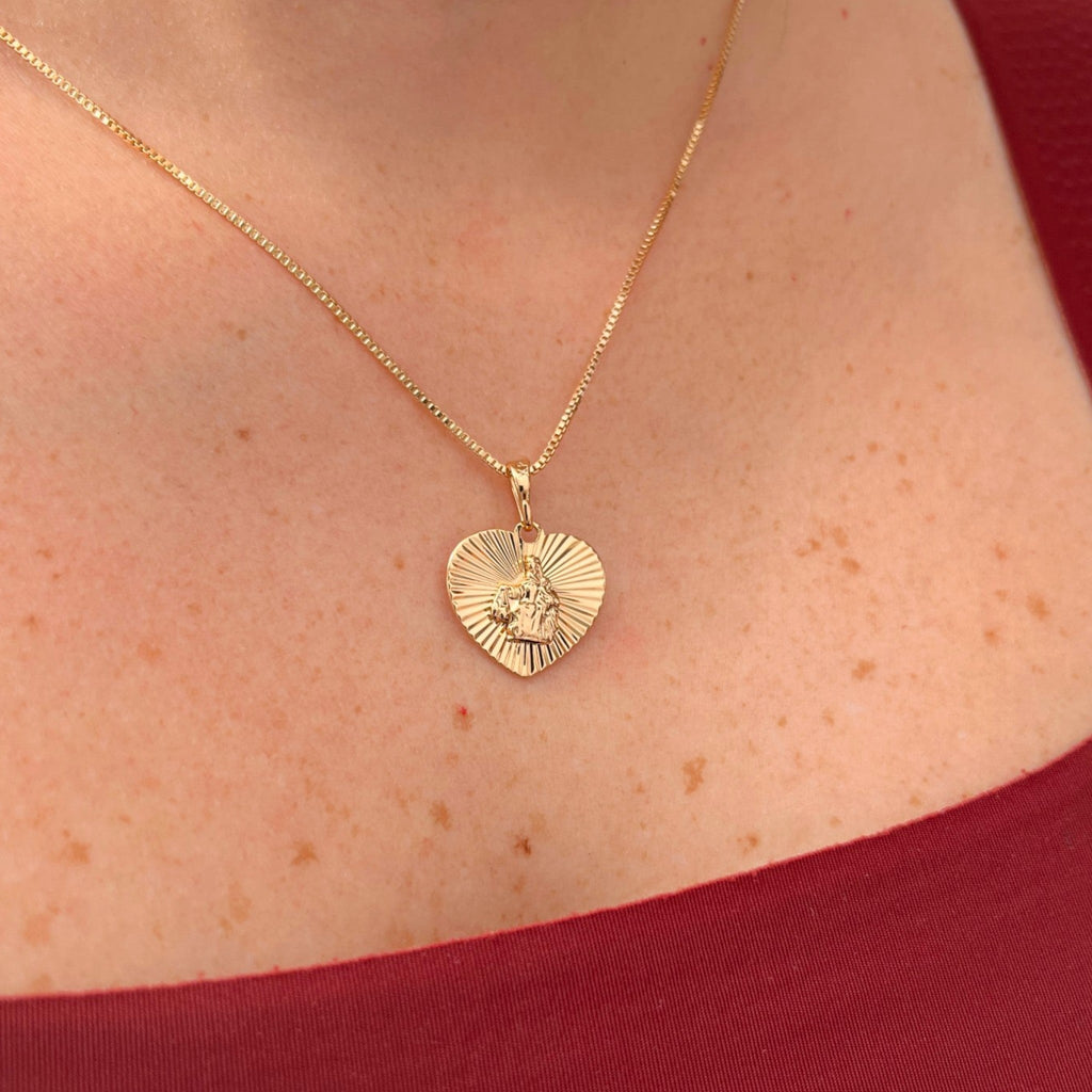 Cadena de tejido Veneciana en acabado dorado, acompañada por un dije en forma de corazón doble cara o reversible. Por una cara presenta la imagen del Sagrado Corazón de Jesús con halo radiante y por la otra la figura de la Virgen del Carmen sobre un fondo texturizado de destellos. Una pieza versátil de gran devoción y protección.