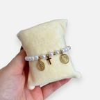 Pulsera de perlas Hv Hermanas Villegas con cruz y medallas religiosas en enchapado oro rosé sobre cojín de exhibición