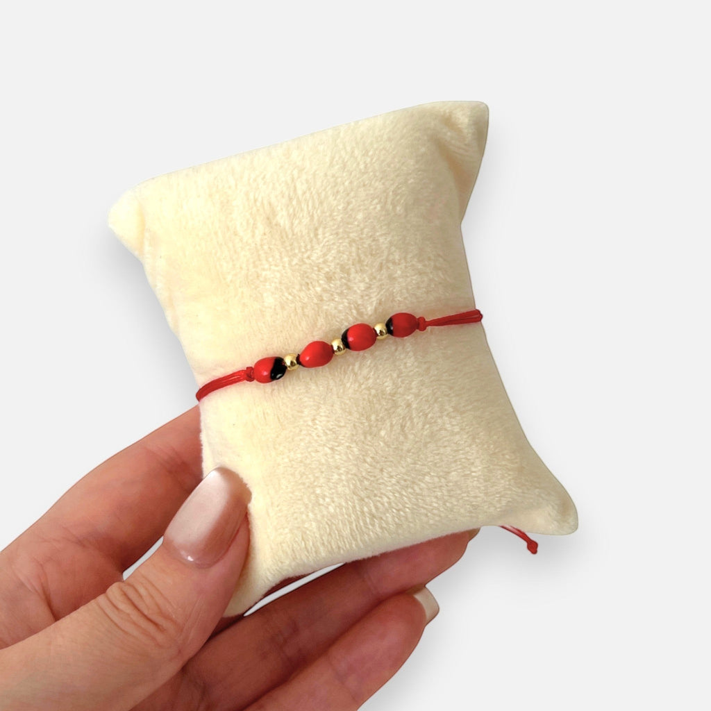 Pulsera Hv Hermanas Villegas de hilo rojo con semillas de huayruro y balines de acero dorado sobre cojín protector
