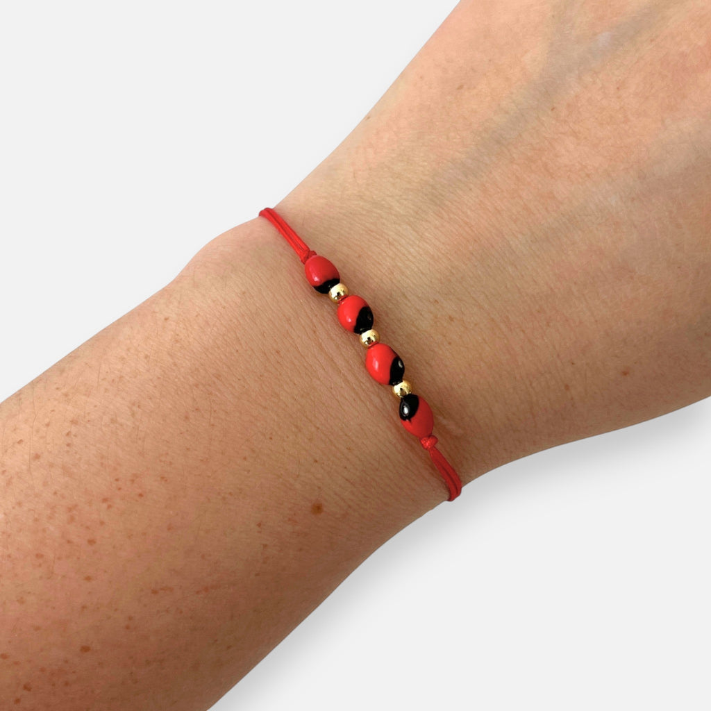Pulsera Hv Hermanas Villegas de hilo rojo con semillas de huayruro y balines de acero dorado en muñeca