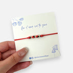Pulsera Hv Hermanas Villegas de hilo rojo con semillas de huayruro y balines de acero dorado sobre tarjeta de regalo