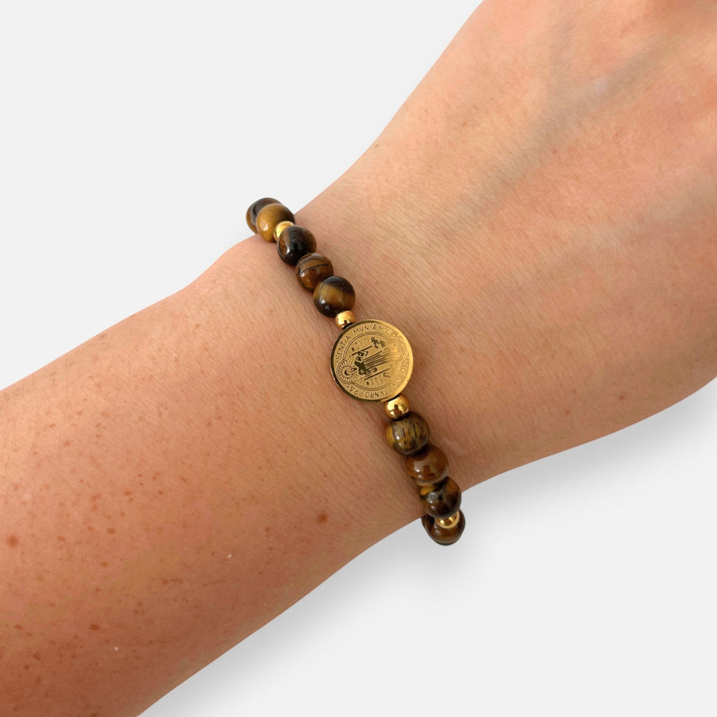 Pulsera Hv Hermanas Villegas de hilo con piedras ojo de tigre y medalla de San Benito en acero dorado en una muñeca
