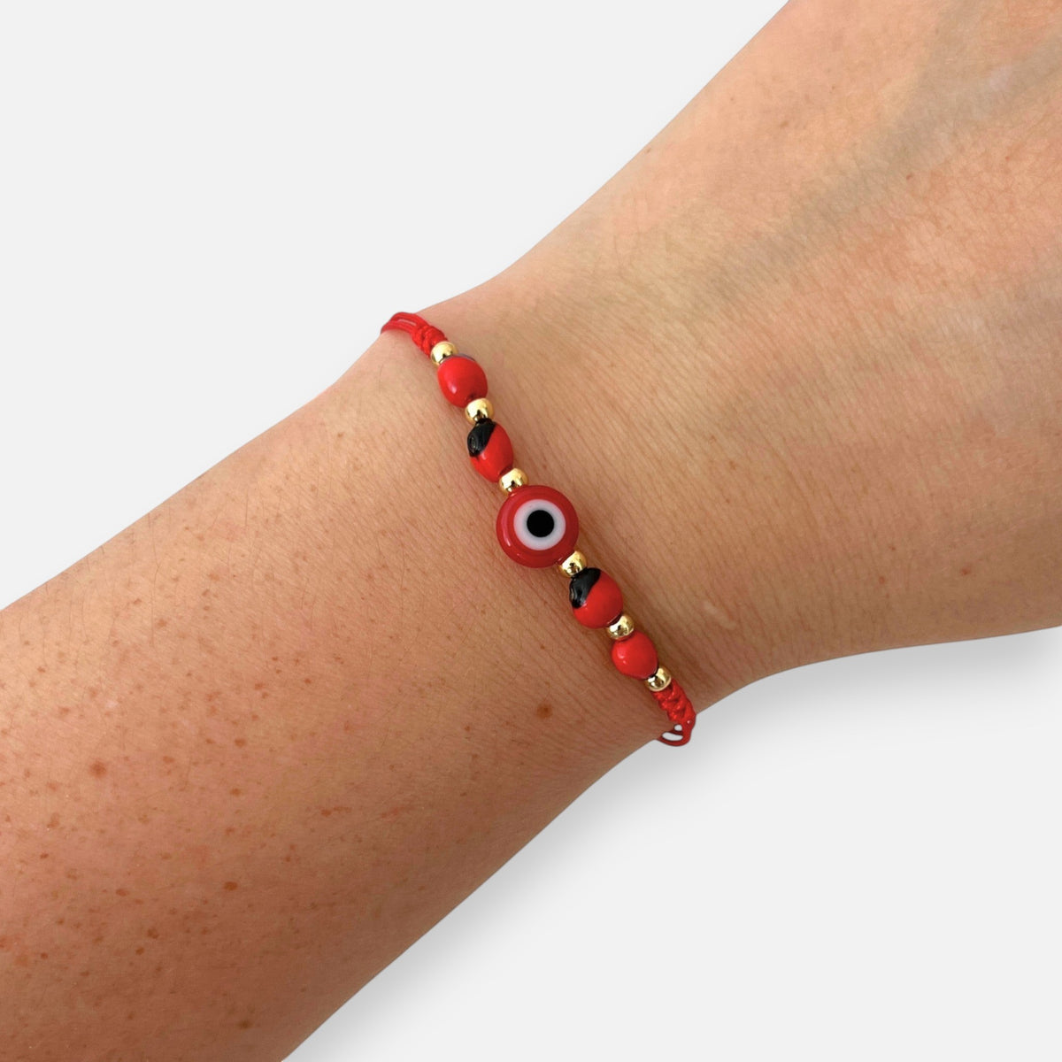 Pulsera Hv Hermanas Villegas de hilo rojo con ojo turco, semillas de huayruro y detalles en acero inoxidable dorado.