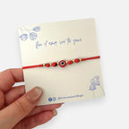Pulsera Hv Hermanas Villegas de hilo rojo con ojo turco, semillas de huayruro y detalles en acero inoxidable dorado.