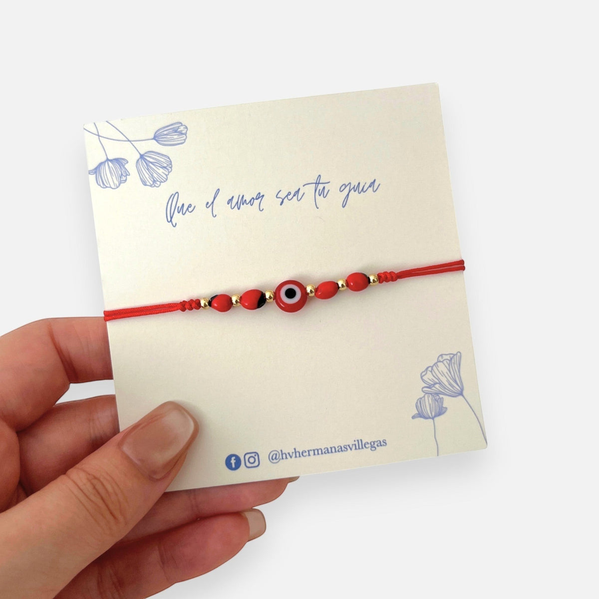 Pulsera Hv Hermanas Villegas de hilo rojo con ojo turco, semillas de huayruro y detalles en acero inoxidable dorado.