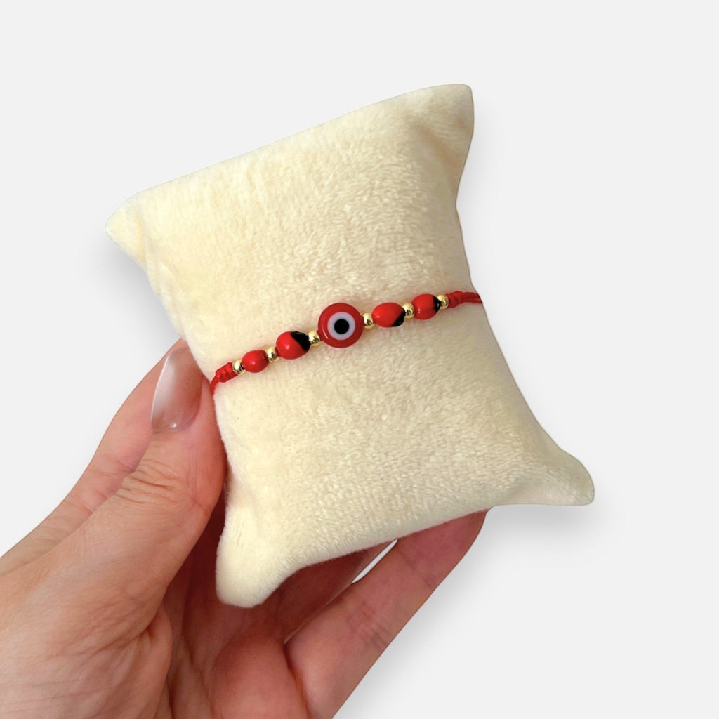 Pulsera Hv Hermanas Villegas de hilo rojo con ojo turco, semillas de huayruro y detalles en acero inoxidable dorado.