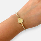 Pulsera Hv Hermanas Villegas de hilo con cuentas de Murano crema, dije de San Benito en acero dorado y circones.