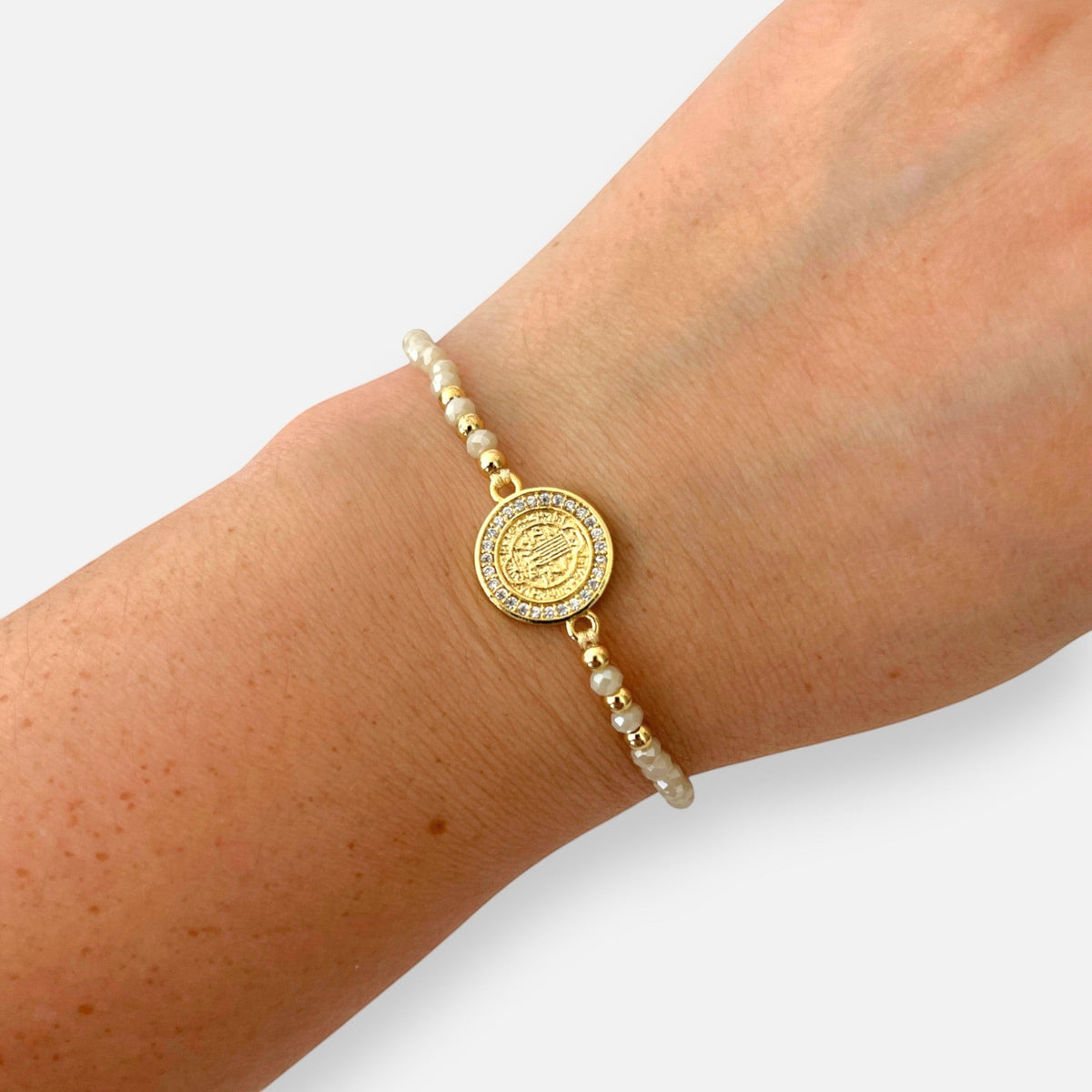 Pulsera Hv Hermanas Villegas de hilo con cuentas de Murano crema, dije de San Benito en acero dorado y circones.