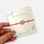 Pulsera Hv Hermanas Villegas de hilo rojo con medalla de San Benito, circones brillantes y detalles en acero dorado.