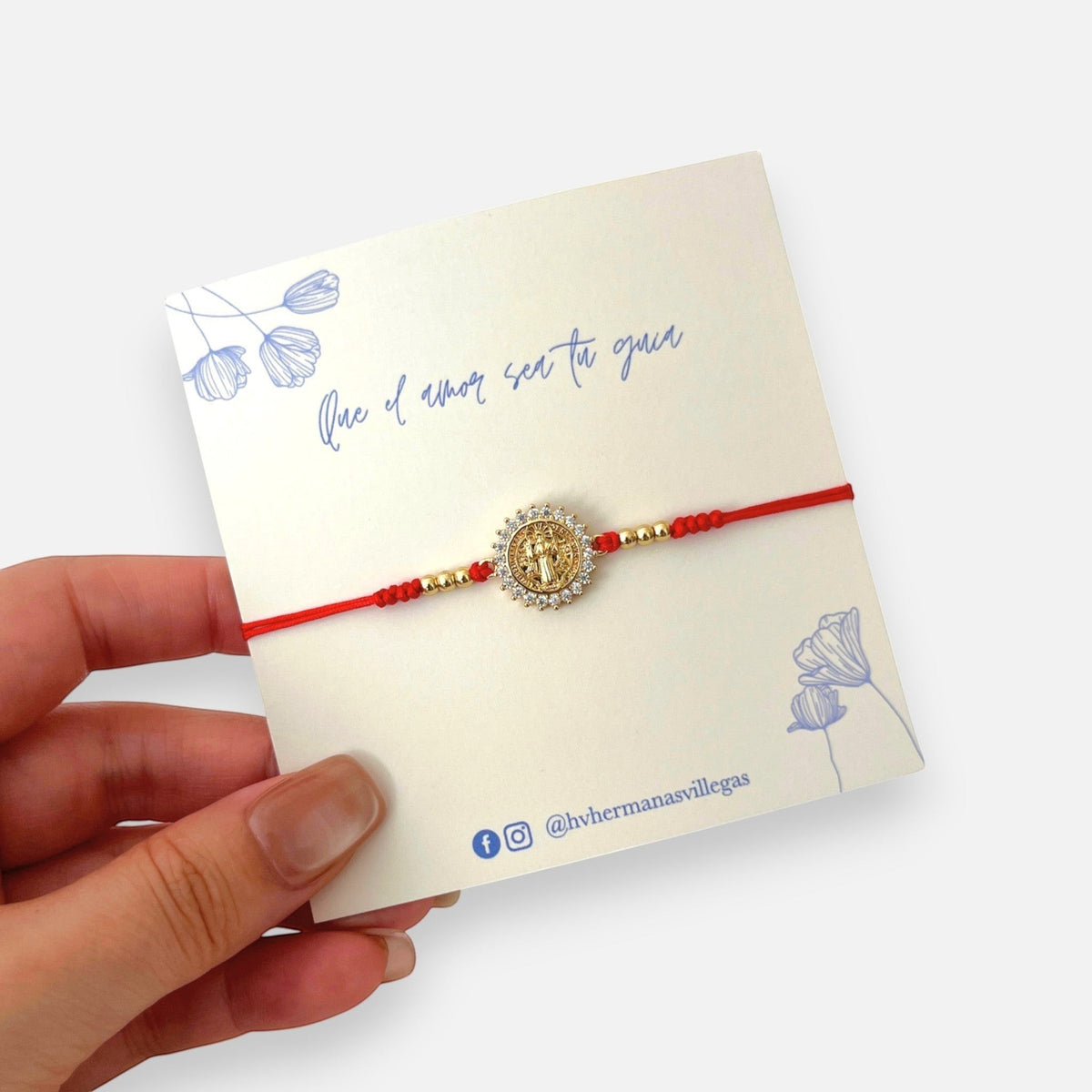Pulsera Hv Hermanas Villegas de hilo rojo con medalla de San Benito, circones brillantes y detalles en acero dorado.