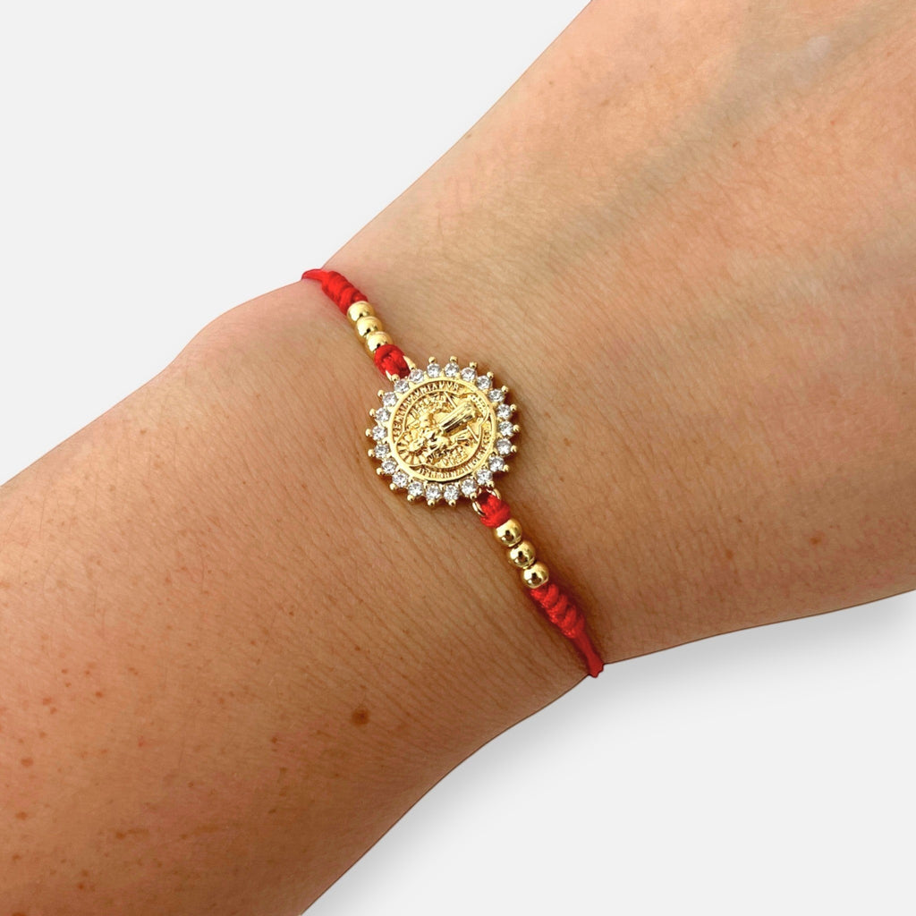 Pulsera Hv Hermanas Villegas de hilo rojo con medalla de San Benito, circones brillantes y detalles en acero dorado.