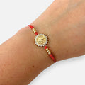 Pulsera Hv Hermanas Villegas de hilo rojo con medalla de San Benito, circones brillantes y detalles en acero dorado.