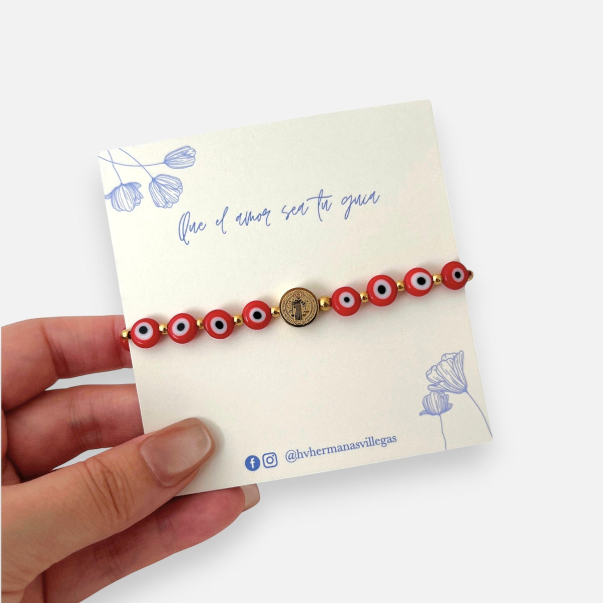 Pulsera Hv Hermanas Villegas de hilo rojo con ojos turcos y medalla de San Benito en acero dorado sobre tarjeta