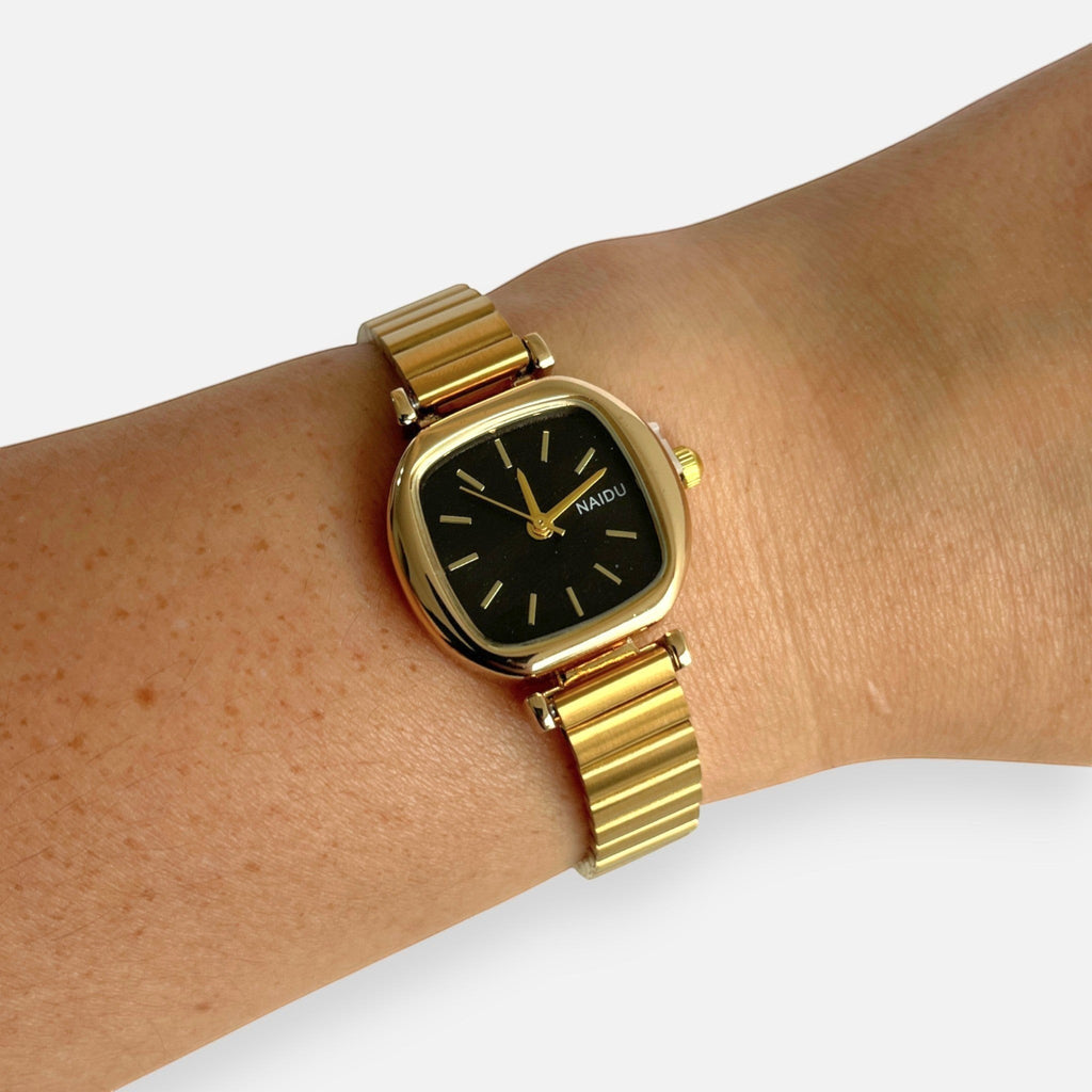 Reloj Hv Hermanas Villegas con caja cuadrada dorada, dial negro y correa extensible de acero en muñeca femenina