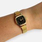 Reloj Hv Hermanas Villegas con caja cuadrada dorada, dial negro y correa extensible de acero en muñeca femenina
