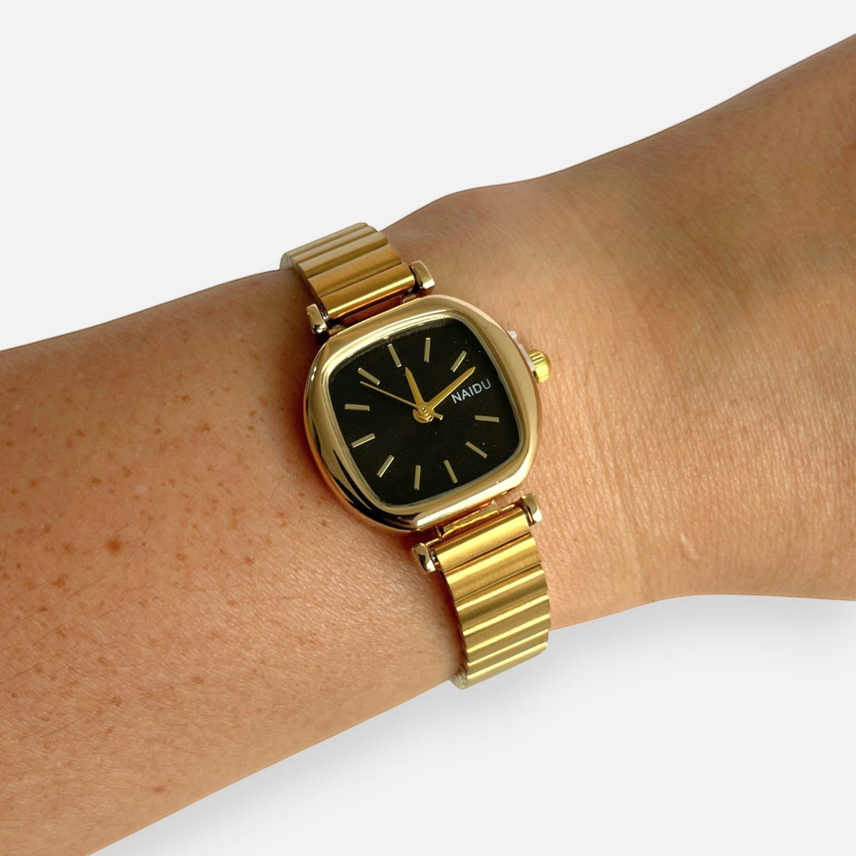 Reloj Hv Hermanas Villegas con caja cuadrada dorada, dial negro y correa extensible de acero en muñeca femenina