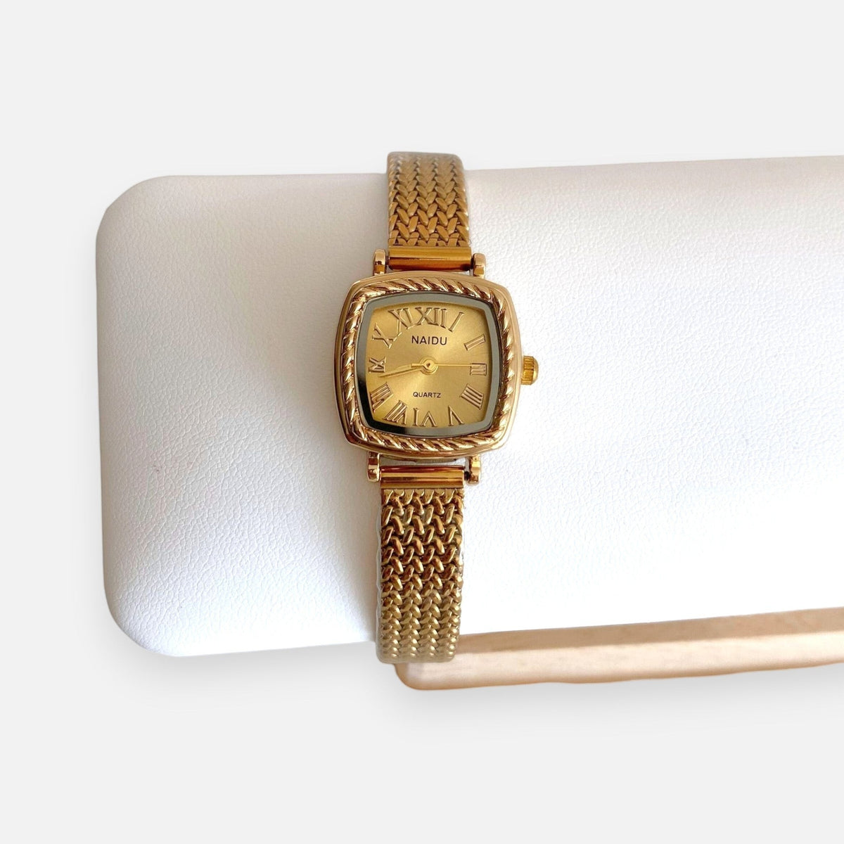 Reloj analógico Hv Hermanas Villegas con caja cuadrada dorada, dial con números romanos y correa de acero trenzada.