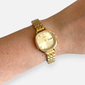 Reloj Hv Hermanas Villegas cuadrado de acero dorado con dial champaña, correa de eslabones y calendario en muñeca