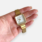 Reloj analógico Hv Hermanas Villegas con caja cuadrada dorada, dial blanco y correa metálica extensible estilo vintage.