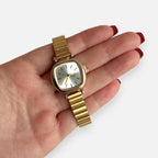 Reloj Hv Hermanas Villegas cuadrado dorado con dial champán y correa extensible flexible en mano femenina