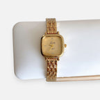 Reloj analógico Hv Hermanas Villegas de acero dorado con caja cuadrada, dial texturizado y detalles brillantes