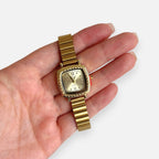 Reloj Hv Hermanas Villegas cuadrado dorado con bisel labrado, correa de eslabones y dial con números romanos.