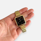 Reloj Hv Hermanas Villegas con caja cuadrada dorada, dial negro minimalista y correa metálica extensible en mano
