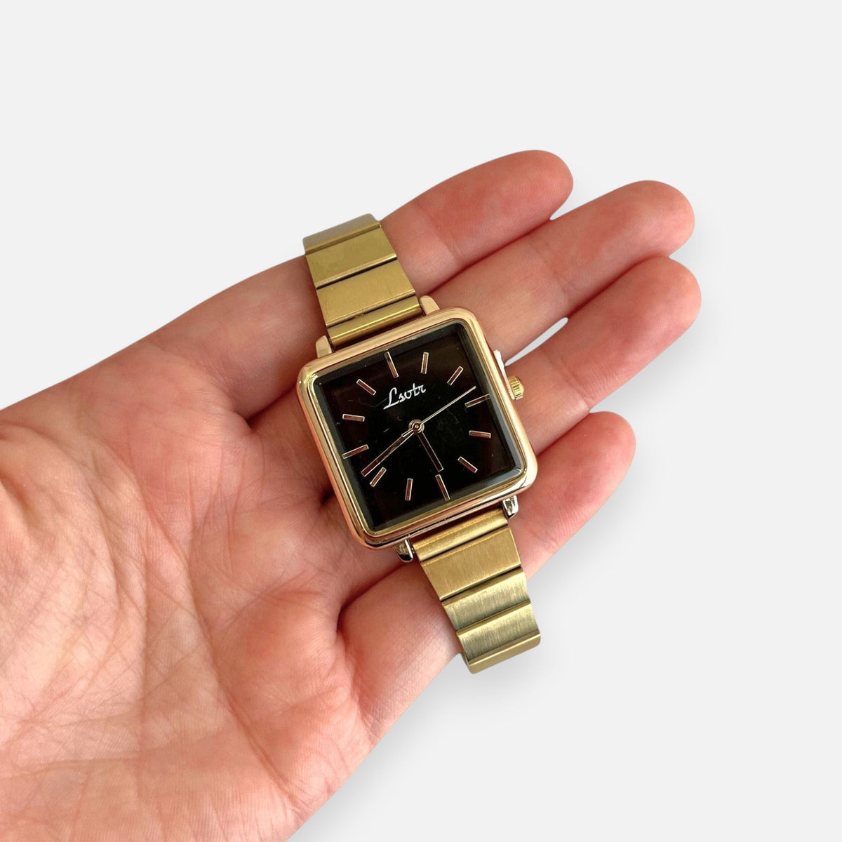 Reloj Hv Hermanas Villegas con caja cuadrada dorada, dial negro minimalista y correa metálica extensible en mano