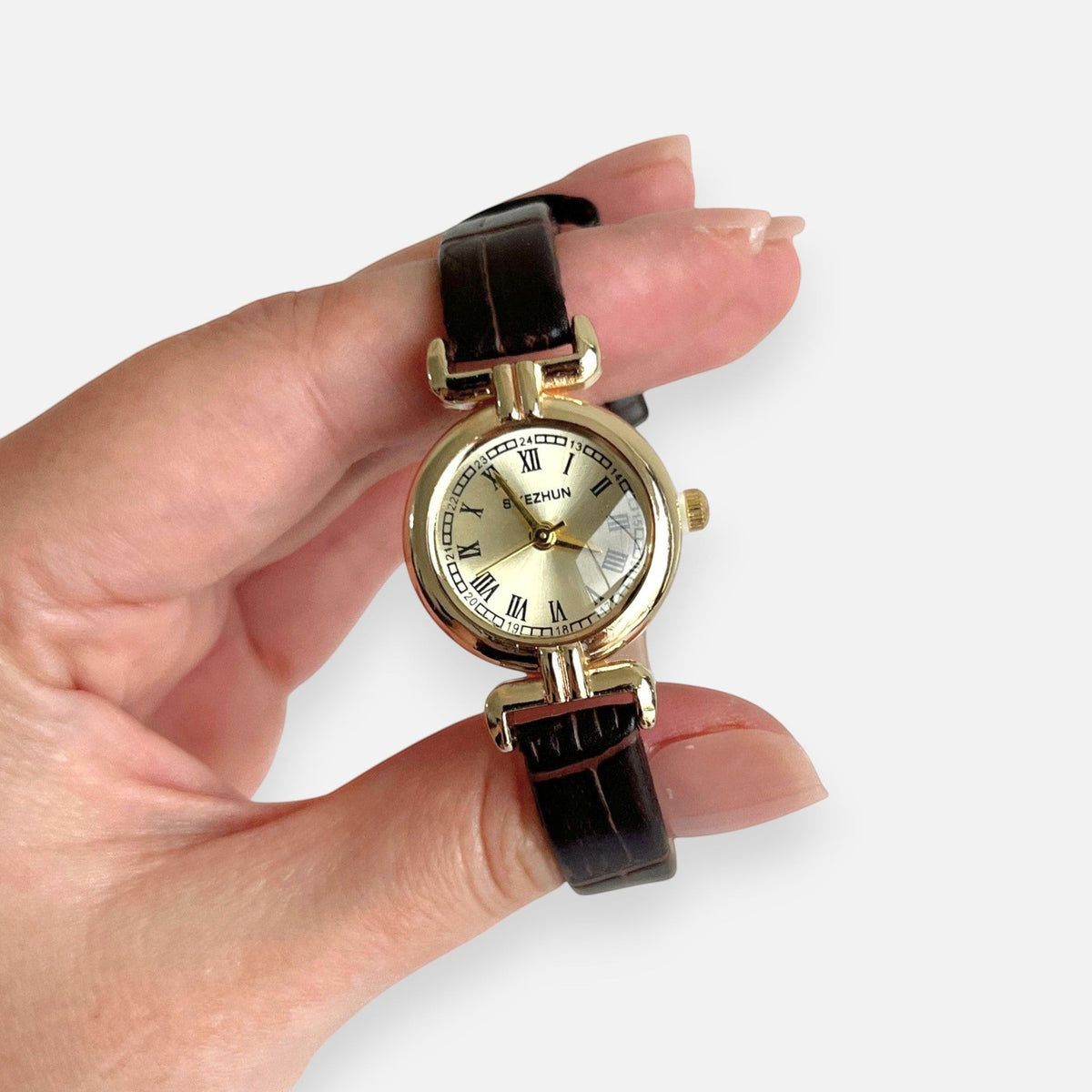 Reloj Hv Hermanas Villegas con caja redonda dorada, dial champán, números romanos y correa de cuero vegano marrón.