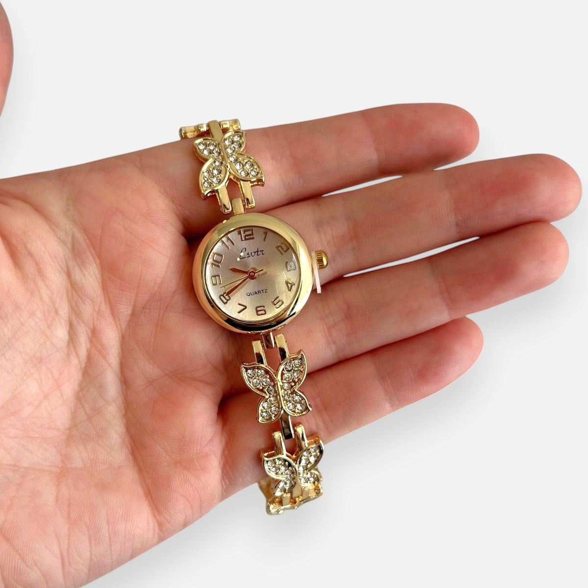 Reloj analógico Hv Hermanas Villegas dorado con mariposas brillantes y dial claro en una mano
