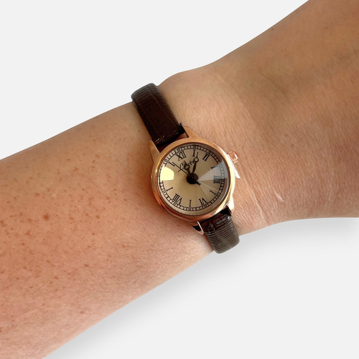 Reloj Hv Hermanas Villegas de oro rosa con dial champán, números romanos y correa de cuero vegano marrón oscuro.