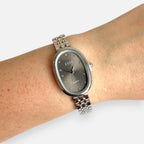 Reloj Hv Hermanas Villegas con caja ovalada plateada, dial gris topo y correa de acero inoxidable en una muñeca