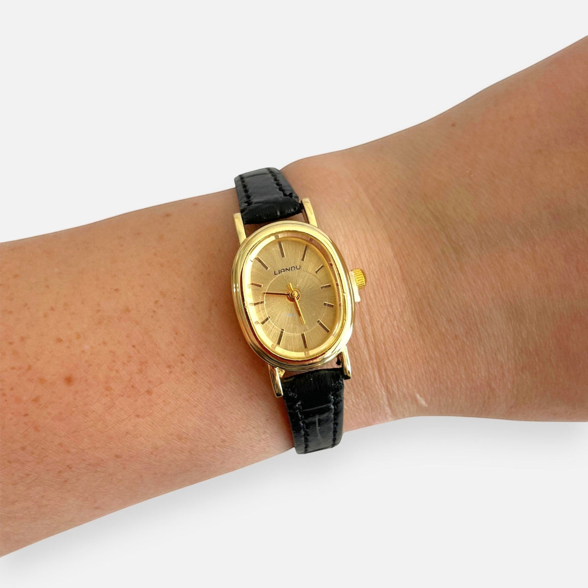 Reloj Hv Hermanas Villegas ovalado dorado con dial cepillado y correa de cuero vegano negro en la muñeca