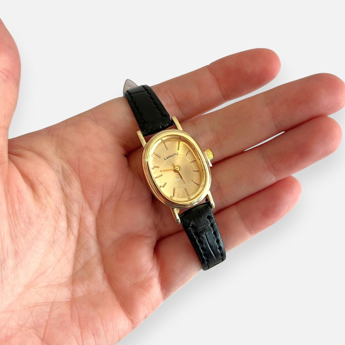 Reloj Hv Hermanas Villegas con caja ovalada dorada, dial cepillado y correa de cuero vegano negro en una mano