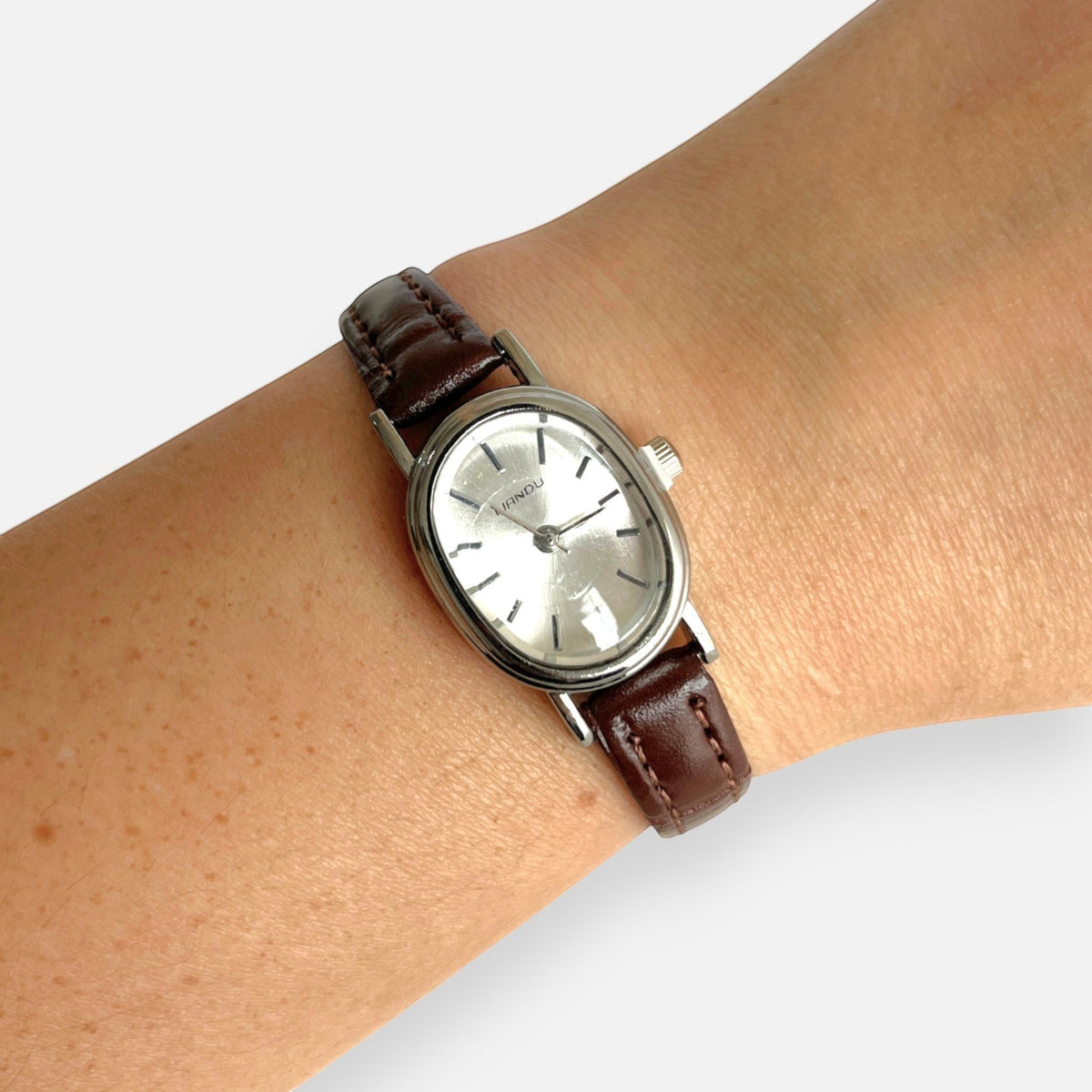 Reloj Hv Hermanas Villegas con caja ovalada plateada, dial satinado y correa de cuero vegano café en muñeca femenina.