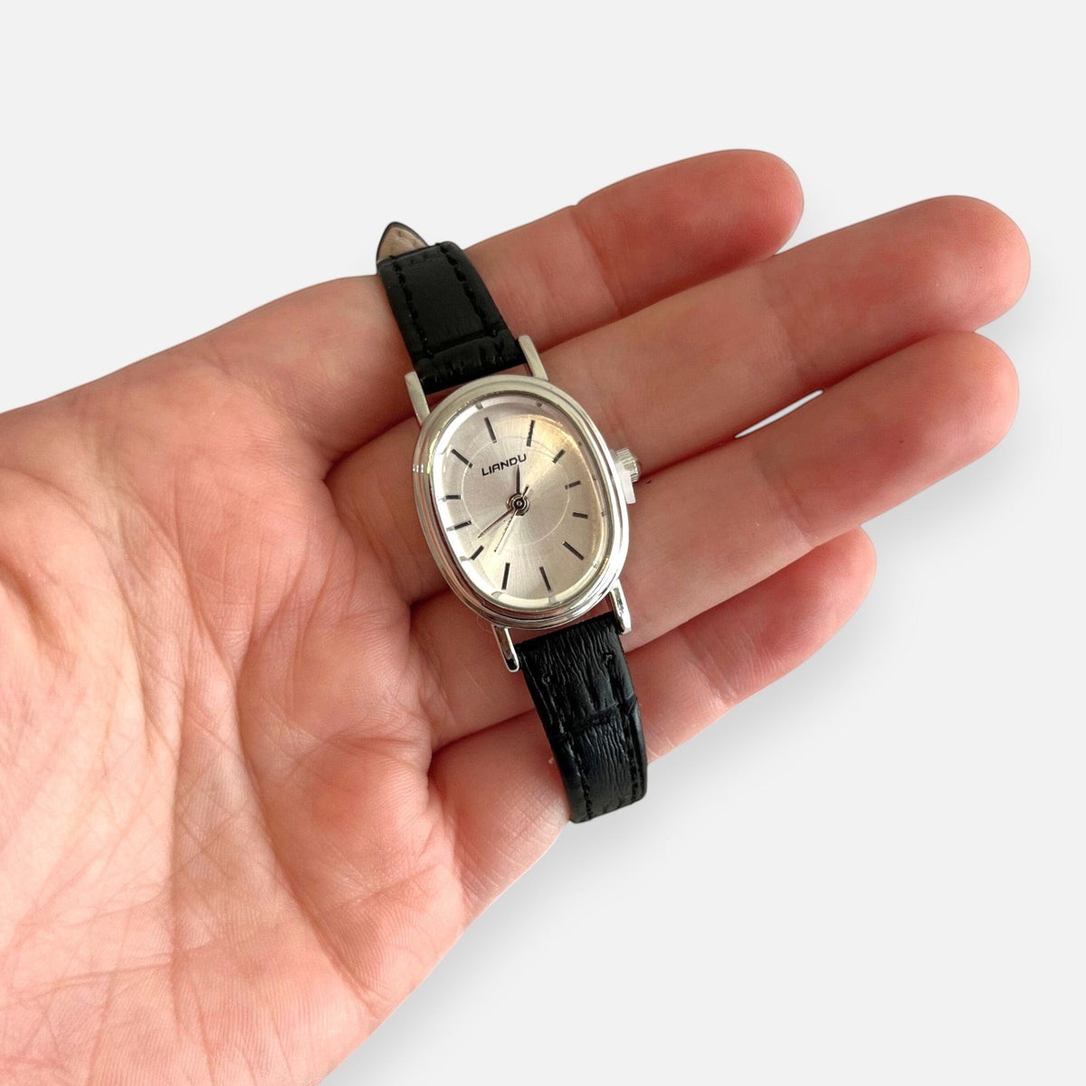 Reloj Hv Hermanas Villegas con caja ovalada plateada, dial satinado y correa de cuero vegano negro en una mano